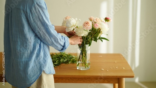 Woman arranging peony and rose flowers in a glass vase, creating a beautiful bouquet for home decoration, enjoying a peaceful floristry hobby activity in soft daylight