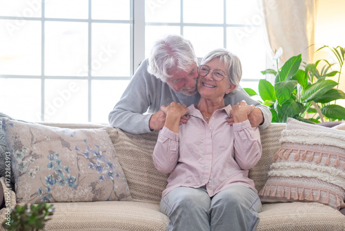 Happy senior couple sharing an affectionate moment at home. Elderly man kissing his wife sitting on a cozy sofa in living room. Concept of love, romance, affection, happy retirement
