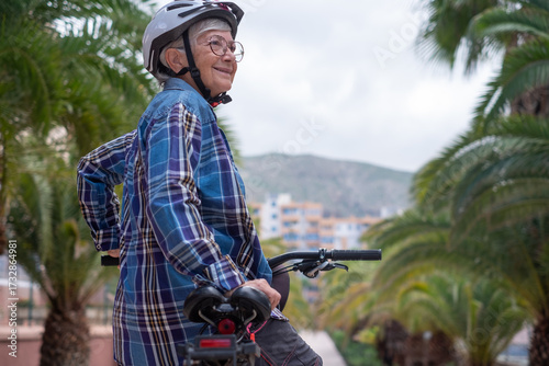 Smiling senior caucasian cyclist woman in leisure activity in urban park riding with electric bike enjoying healthy lifestyle