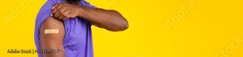 Covid-19 And Flu Vaccination. African American Guy Vaccinated With Antiviral Vaccine Shot Posing Rolling Up Sleeve Showing Arm With Medical Plaster On Yellow Background. Coronavirus Prevention