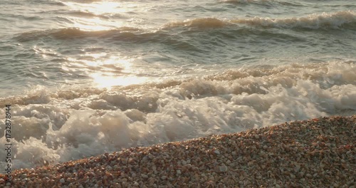 Calm sea waves crash against a quiet pebble beach during a warm sunset. The golden sun reflects off the surface of the water, creating a peaceful and serene natural backdrop.
