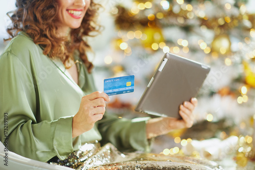 A happy woman with curly brown hair holds a credit card and tablet, engaged in online holiday shopping. Soft bokeh lights in the blurred background add to the warm, celebratory feel.