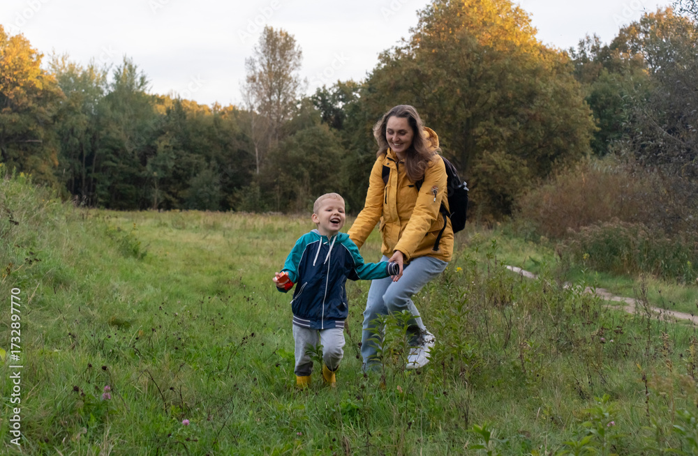 Obraz premium Joyful mother and laughing child running hand in hand through autumn meadow with golden trees. Concept of family bonding and healthy outdoor lifestyle with children.