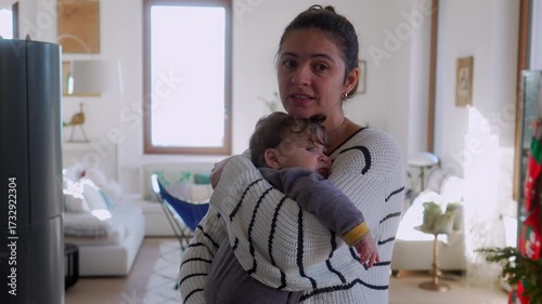 Mother gently holding tired baby close to chest while comforting child in warm embrace inside cozy home living room with window light and Christmas tree