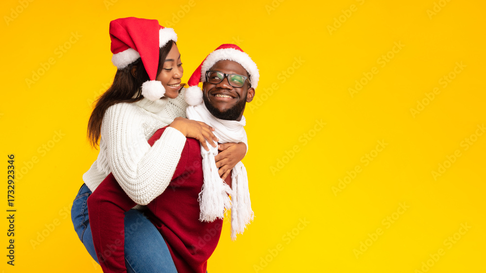Fototapeta premium Xmas Concept. African American Husband Carrying Wife Piggyback Wearing Santa Hats On Christmas Posing Over Yellow Studio Background. Spouses Having Fun Celebrating New Year Holiday