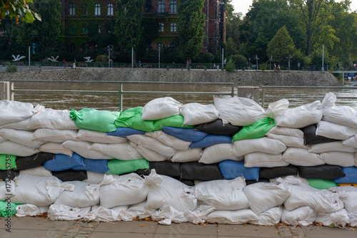 Multiple layers of colorful sandbags stacked as flood protection barrier along urban riverfront with historic ivy-covered buildings. Comprehensive flood defense and emergency preparedness measures.