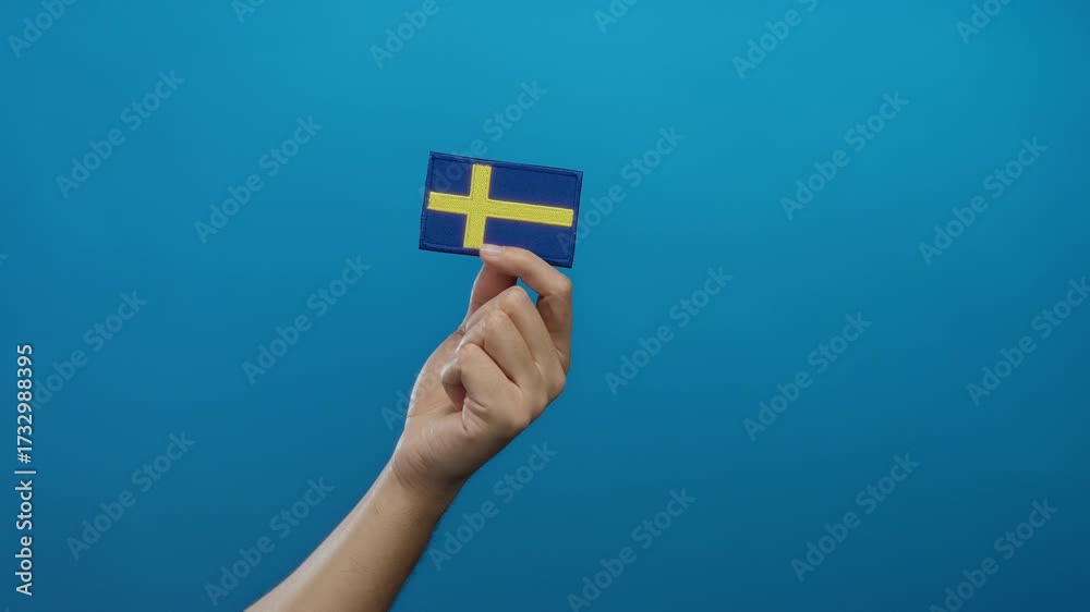 Man holding swedish flag embroidery against blue isolated background, showcasing national identity with focus on hand detail and rich cultural symbolism.