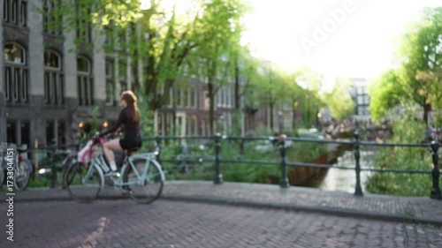 Wallpaper Mural Woman cycling along amsterdam canal with blurred, daylight background of historic buildings and trees, capturing serene urban life and european architecture vibe. Torontodigital.ca