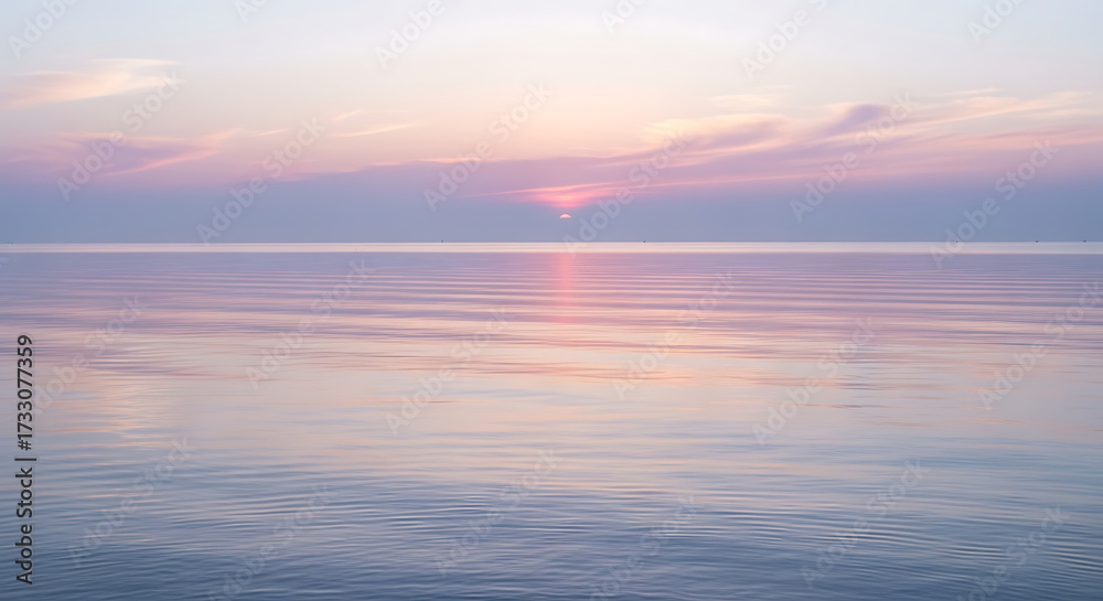 Fototapeta premium Calm ocean water reflects the soft colors of a pastel sunset, with the sun partially obscured by clouds on the horizon.