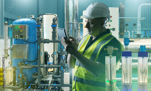 Engineer in safety vest and helmet using tablet to check industrial equipment, pipelines and gauges in factory, monitoring and maintenance. Industry.