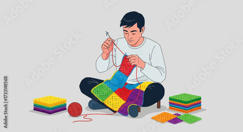 Man sitting on floor knitting a colorful blanket with yarn and needles.