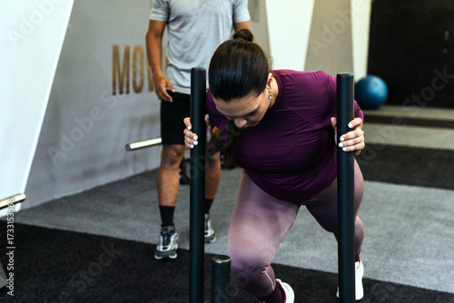 Latin american woman intensively sled pushing, training strength and endurance with her personal trainer and coach in a modern gym