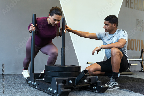 Latin american woman intensely pushing a weighted sled in a gym, receiving guidance from her male personal trainer