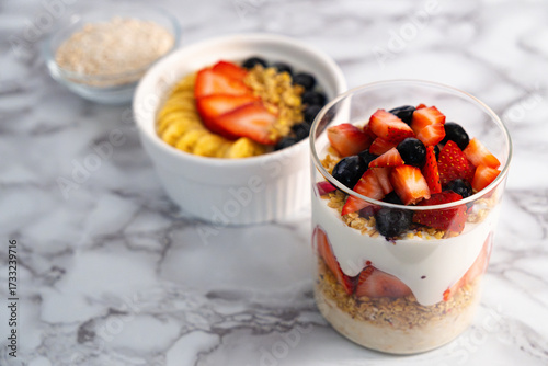
Clean and simple photography of a healthy yogurt dessert made with banana, strawberries, and oats placed on a clear surface showing 3 containers.