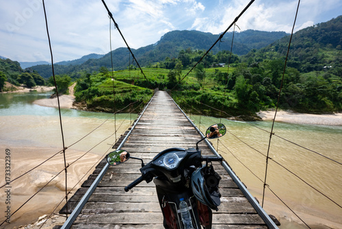 Obraz na plátně An adventure travel shot of a motorcycle parked on a rustic wooden suspension bridge spanning across a flowing river in the scenic mountains of northern Vietnam