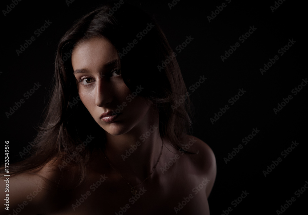 Fototapeta premium Studio portrait of young woman with long brown hair and bare shoulders, highlighted by dramatic lighting against a black background