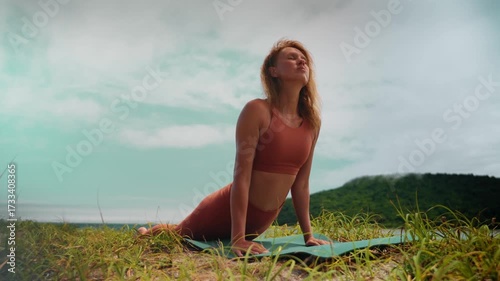 Female yogi in red outfit stretches in upward dog pose on grass by ocean. Calmness, discipline, and outdoor energy highlight meditation practice and natural wellness lifestyle.