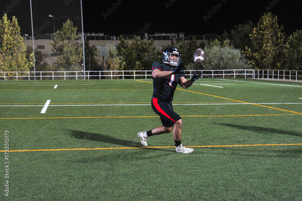 Fototapeta premium Male athlete in uniform playing american football, actively catching the ball during night game on green field