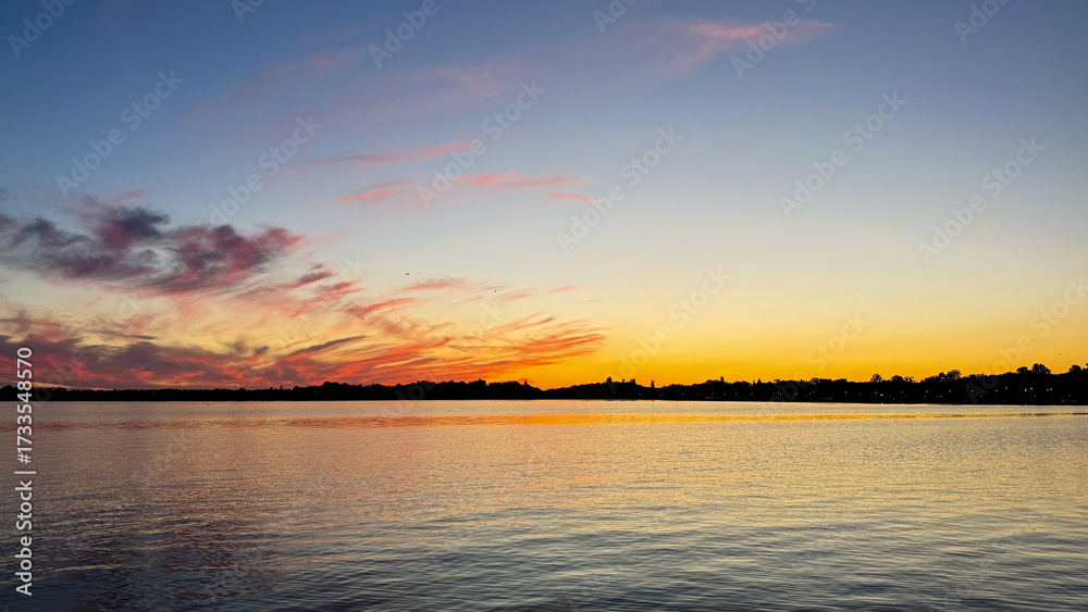 Fototapeta premium Sunset paints the sky in swirling tangerine and indigo, reflecting tranquility on water, invoking Serenity Day, Midsummer Festival