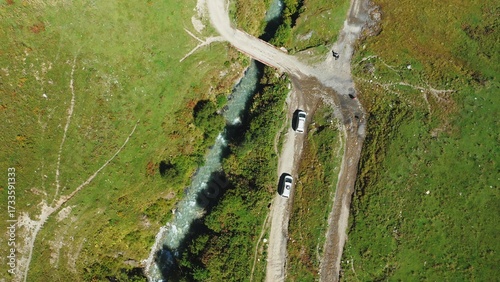 A top-down aerial view of two cars driving along a dirt road along a mountain river to the bridge.