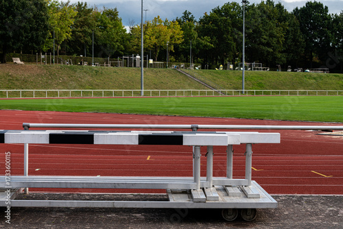 A track field features hurdles placed on the red running surface. In the background, lush green grass is visible, surrounded by trees and sports facilities. The scene is calm and clear.