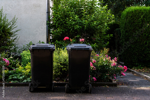 Two black trash bins stand on a path surrounded by vibrant flowers and greenery. The scene is set in a peaceful residential area, showcasing a blend of nature and daily life.