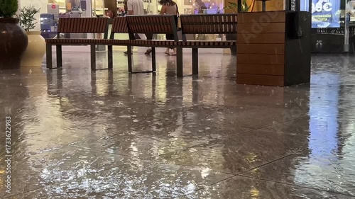 Outdoor benches in a public area, seen on a rainy day. People late shoppers running under rain on background. wet floor reflecting lights from nearby shops