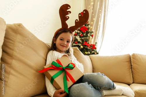Happy little girl in reindeer antlers holding wrapped Christmas gift with ribbon, sitting on beige sofa near decorated tree with lights. Cozy holiday atmosphere at home.