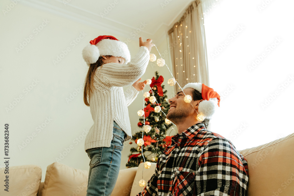 Obraz premium Father and daughter decorating for Christmas together, both wearing Santa hats, smiling and playing with glowing string lights at home near a Christmas tree, capturing joy, love, and family bonding.