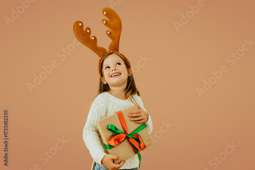 Overjoyed little girl in reindeer antlers holding Christmas gift with ribbon, wearing white sweater and jeans. Festive child portrait on warm background celebrating holiday season.