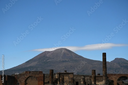 Broken columns and arches from the Forum in Pompeii are seen with Mount Vesuvius in the background. A road is visible winding up the mountain. The sky is blue with a few clouds.