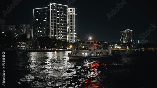 A large motor ship sails along a river in a night city. Reflections and light of buildings are visible on the water. Shooting in motion