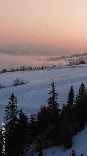 Aerial drone view of warm winter foggy dawn over rolling snow-capped mountains and pine forest. Beauty in nature and calmness