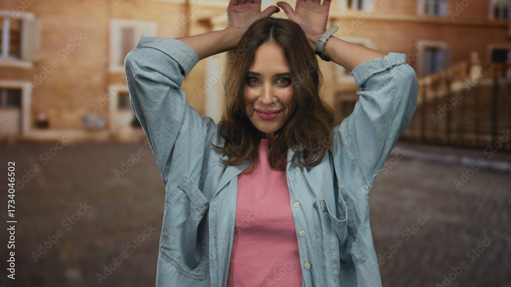 Fototapeta premium Young hispanic woman in casual denim and pink attire playfully makes ear gesture on a bustling city street, exuding joy and warmth in an engaging urban setting.