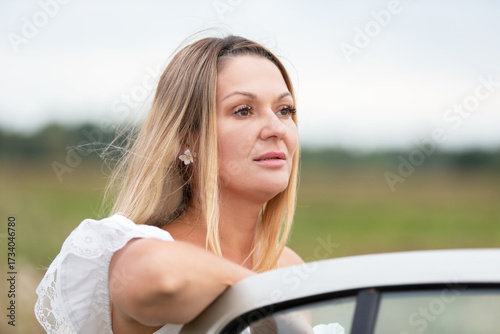 Woman with long hair leans out of car window in a green field under a cloudy sky during daylight hours