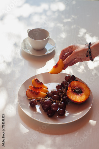 Plate of Grapes and Peach with Hand Holding Peach Slice and Coffee Cup on White Table, Sardinia