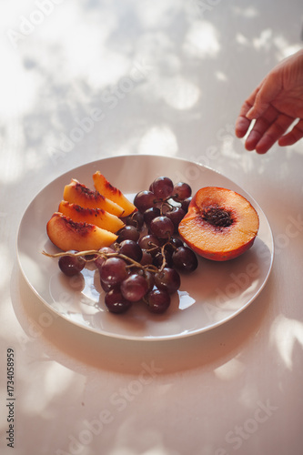Female Hand Reaching for Peach and Grapes on White Plate, Sardinia