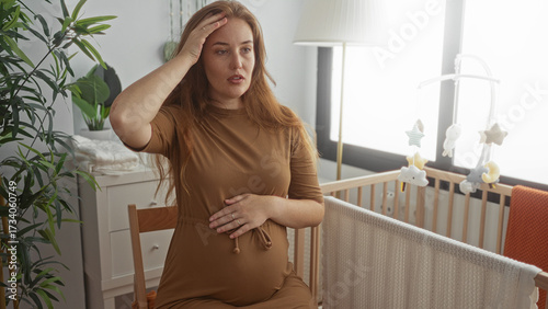 фотографія Young caucasian woman holding her pregnant belly with one hand while touching her temple in studio; maternal discomfort