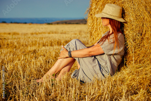 red-haired beautiful woman with hat in field with haystacks against blue sky