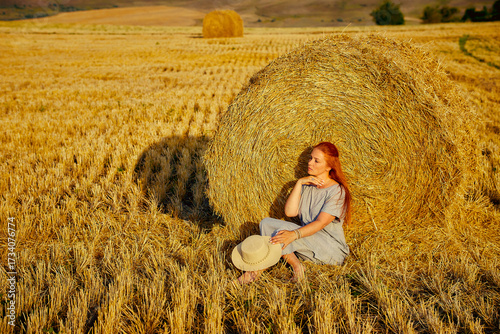 red-haired beautiful woman with hat in field with haystacks against blue sky