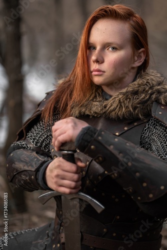 Red-haired warrior stands resolute amidst a wintry forest, clad in armor and gripping a sword.
