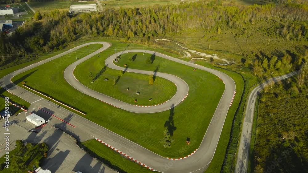 Aerial drone footage orbiting and flying around a go kart or karting racing speedway track in a sunny summer or autumn countryside. The track is in the middle of a forest and besides a highway road.