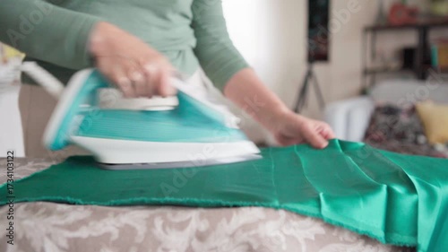 4K shot of woman's hands using a steam iron to smooth wrinkles from green fabric dress. This domestic routine captures  repetitive action of housework and laundry. 
