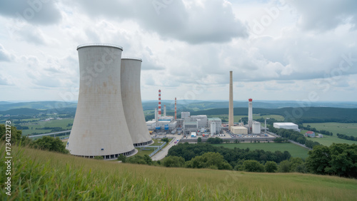 Large cooling towers dominate landscape, surrounded by green hills and power plant. scene conveys industrial energy production and environmental