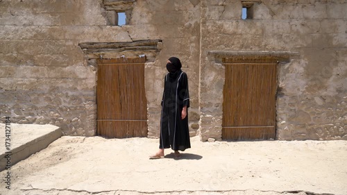 A woman dressed in traditional Arab clothes passes through an archaeological place filled with historical stone structures. The sun shines brightly, illuminating the environment.