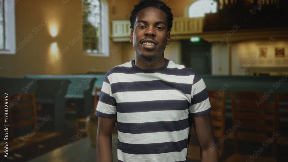 Obraz premium Young african american man in striped shirt gestures thumbs down among wooden pews in church building; disapproval.