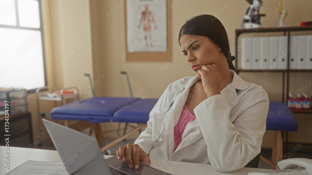 Obraz premium Woman doctor in white coat with hand on chin typing on laptop in clinic building beside anatomy chart; thought concern.