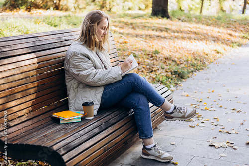 Young woman writing in notebook on bench in autumn park.