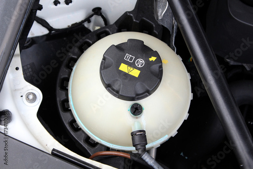 Close-up of modern car coolant reservoir with black cap marked by warning labels and yellow arrows, showing essential engine maintenance component under hood. Car cooling system.