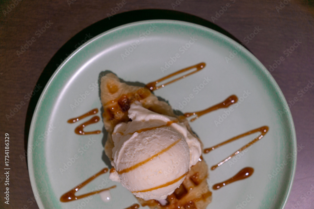 Fototapeta premium A scoop of vanilla ice cream, placed on a waffle, drizzled with caramel sauce, served on a light green plate, set against a dark background